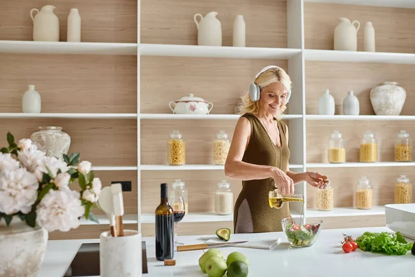 Glückliche Frau mittleren Alters in drahtlosen Kopfhörern, die Salat in der Nähe von Flasche und Glas Rotwein zubereitet — Stockfoto