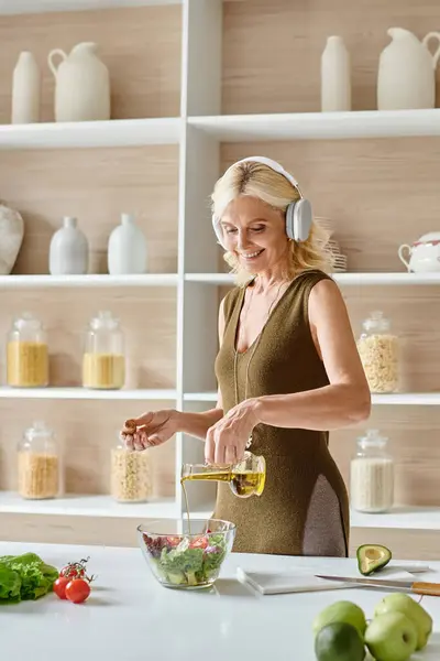 Glückliche Frau mittleren Alters in drahtlosen Kopfhörern, die Salat in der Nähe von frischem Gemüse auf der Arbeitsplatte zubereitet — Stockfoto