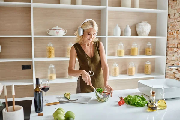 Счастливая женщина средних лет в беспроводных наушниках слушает музыку и делает салат на кухне — стоковое фото