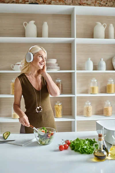 Happy middle aged woman in wireless headphones listening music and making salad in kitchen — Stock Photo