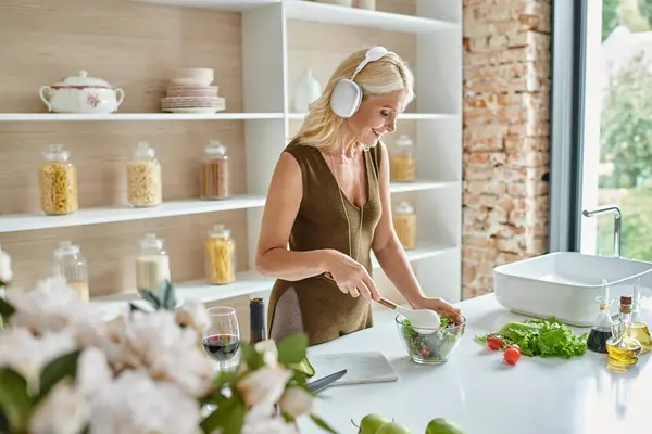 Веселая женщина средних лет в беспроводных наушниках слушает музыку и делает салат на кухне — стоковое фото