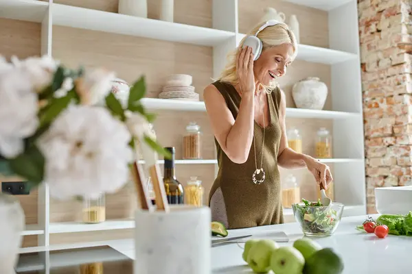 Positive Frau mittleren Alters in drahtlosen Kopfhörern, die Musik hört und Salat in der Küche macht — Stockfoto