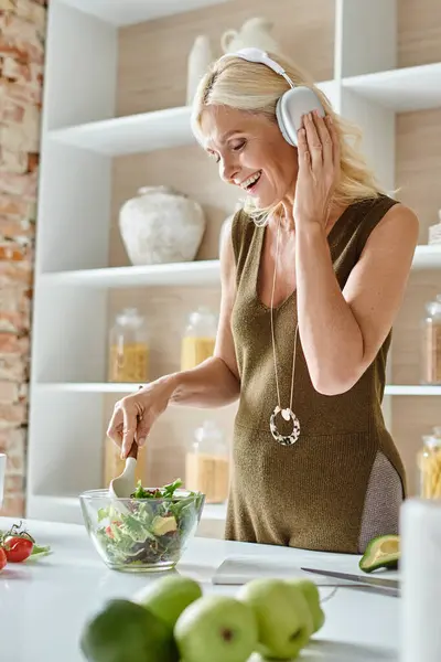 Happy middle aged woman in wireless headphones listening music and mixing salad in kitchen — Stock Photo