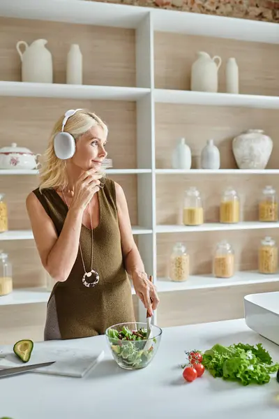 Carefree middle aged woman in wireless headphones listening music and mixing salad in kitchen — Stock Photo