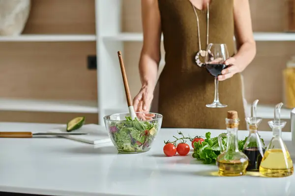 Femme cultivée tenant un verre de vin rouge et debout près de la salade fraîchement préparée dans un bol transparent — Photo de stock