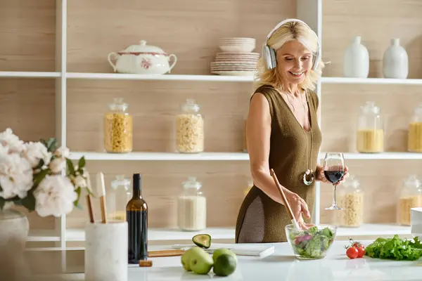 Счастливая женщина средних лет в наушниках, держа бокал вина и стоя возле свежего салата в миске — стоковое фото