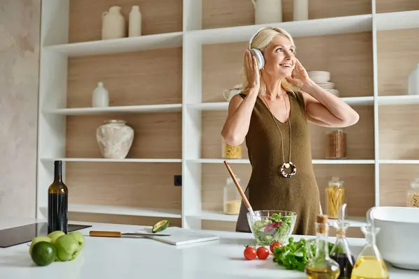Heureuse femme d'âge moyen dans des écouteurs sans fil debout près d'ingrédients frais et bol dans la cuisine — Photo de stock