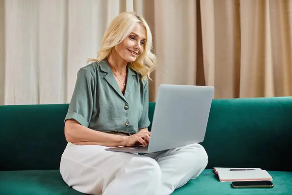 Mulher de meia-idade alegre com cabelo loiro usando laptop enquanto sentado no sofá, trabalho remoto — Fotografia de Stock