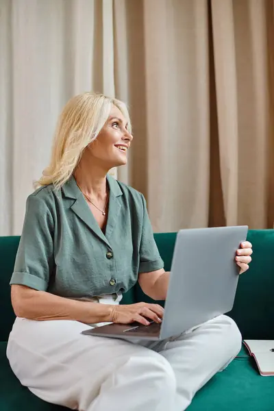 Rayonnante femme d'âge moyen avec des cheveux blonds en utilisant un ordinateur portable tout en étant assis sur le canapé, travail à distance — Photo de stock