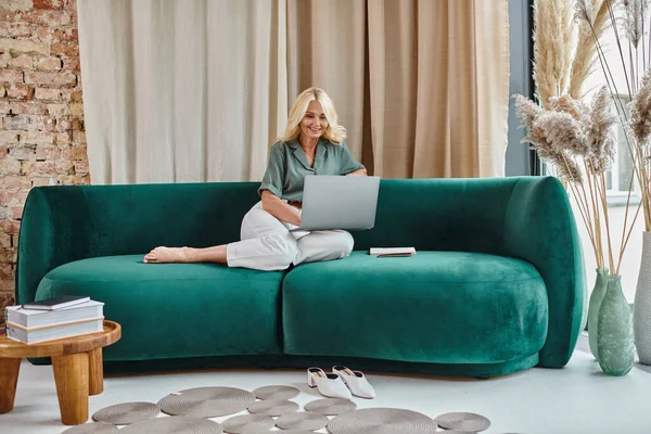Mujer de mediana edad relajada con el pelo rubio usando el ordenador portátil mientras está sentado en el sofá, el trabajo desde casa - foto de stock