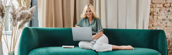 Relaxed middle aged woman with blonde hair using laptop while sitting on sofa, work from home banner — Stock Photo