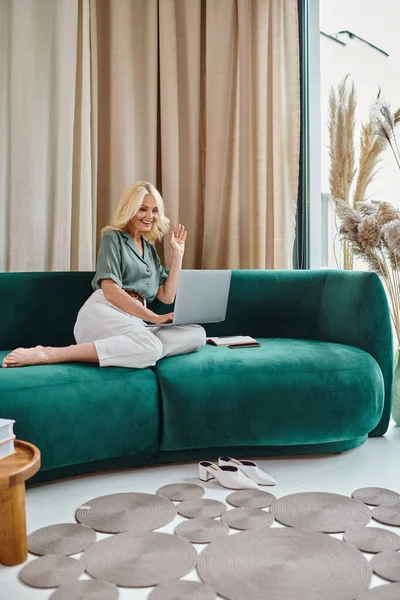 Happy middle aged woman using laptop while sitting on sofa, waving hand during video chat — Stock Photo