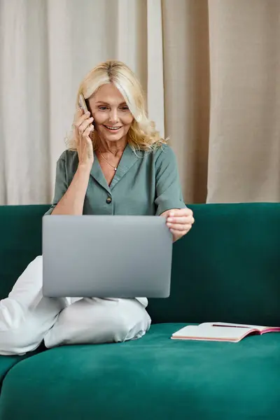 Donna di mezza età con capelli biondi che parla su smartphone e usa il computer portatile, seduta sul divano — Foto stock