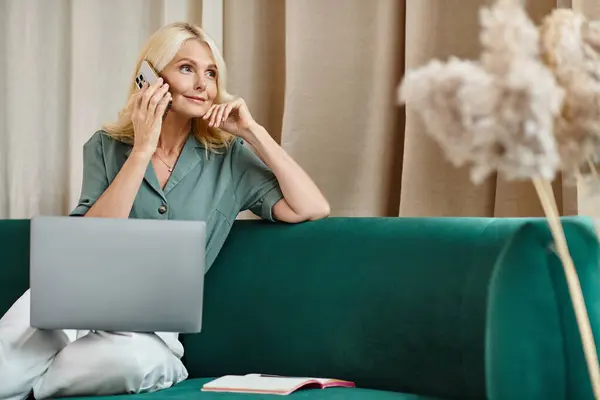 Atractiva mujer de mediana edad con el pelo rubio hablando en el teléfono inteligente y el uso de ordenador portátil en el sofá - foto de stock