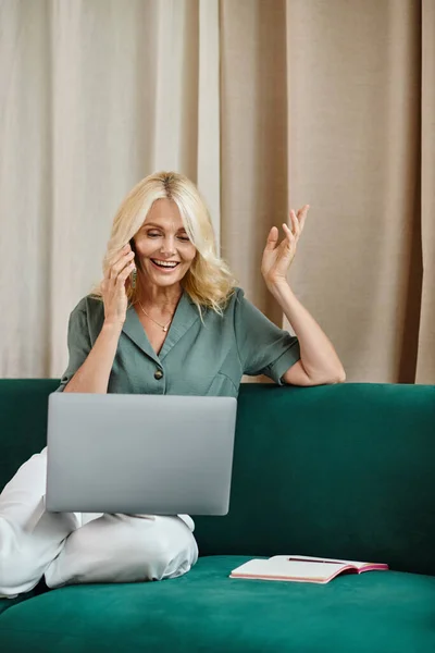Allegra donna di mezza età con i capelli biondi che parla sullo smartphone e utilizza il computer portatile sul divano — Foto stock
