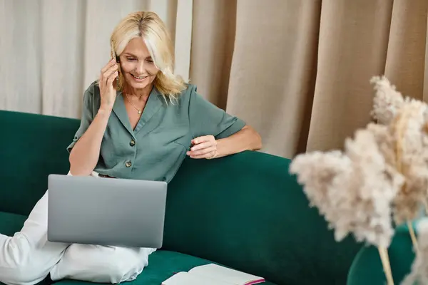 Lächelnde Frau mittleren Alters mit blonden Haaren, die auf dem Smartphone spricht und Laptop im Wohnzimmer benutzt — Stockfoto