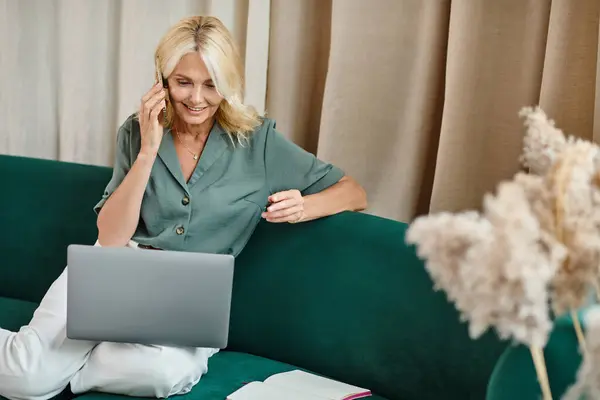 Smiling middle aged woman with blonde hair talking on smartphone and using laptop in living room — Stock Photo