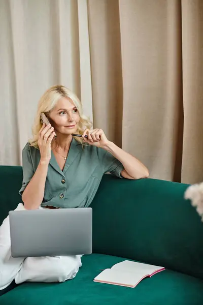 Mujer de mediana edad pensativa con el pelo rubio hablando en el teléfono inteligente y el uso de ordenador portátil en el sofá - foto de stock