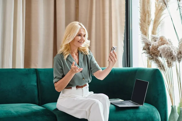 Heureuse femme d'âge moyen ayant appel vidéo sur smartphone, gestuelle et assise sur le canapé près d'un ordinateur portable — Photo de stock