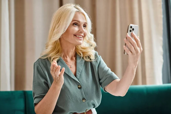 Mujer de mediana edad feliz con el pelo rubio teniendo videollamada en el teléfono inteligente y sonriendo en la sala de estar - foto de stock