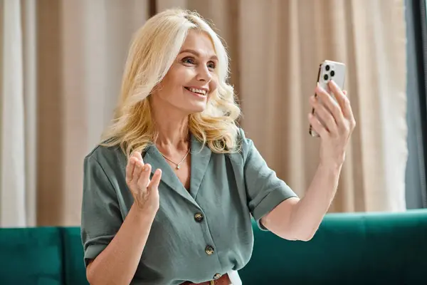 Felice donna di mezza età con i capelli biondi avere videochiamata su smartphone e sorridente in soggiorno — Foto stock