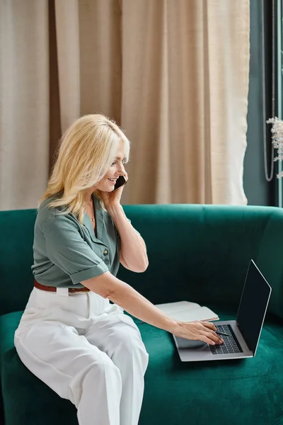 Mujer de mediana edad feliz tener llamada telefónica en el teléfono inteligente y escribir en el ordenador portátil mientras está sentado en el sofá - foto de stock