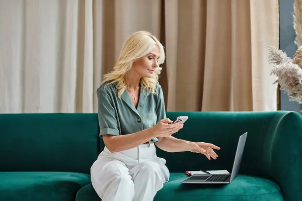 Bionda donna di mezza età utilizzando smartphone e laptop mentre si siede sul divano in soggiorno moderno — Foto stock