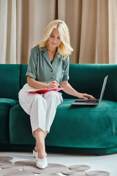 Организованная женщина средних лет с помощью ноутбука и делать заметки, сидя на диване в гостиной — стоковое фото