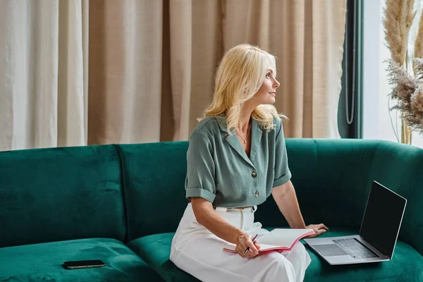 Привлекательная женщина средних лет с помощью ноутбука и делать заметки, сидя на диване в гостиной — стоковое фото