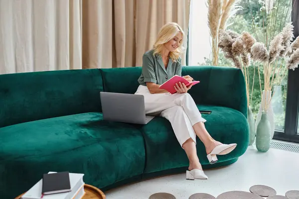 Pleased middle aged woman using laptop and taking notes while sitting on sofa in living room — Stock Photo