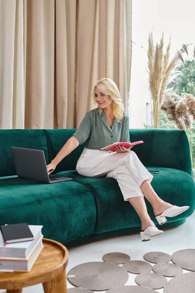 Задоволена жінка середнього віку використовує ноутбук і тримає блокнот, сидячи на дивані у вітальні — стокове фото