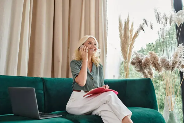 Happy middle aged woman talking on smartphone and taking notes while sitting on sofa with laptop — Stock Photo