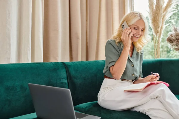 Alegre mujer de mediana edad hablando en el teléfono inteligente y tomando notas mientras está sentado en el sofá con el ordenador portátil - foto de stock