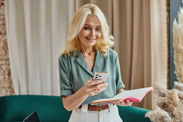 Glückliche Frau mittleren Alters mit blonden Haaren mit Laptop und Notizbuch im modernen Wohnzimmer — Stockfoto