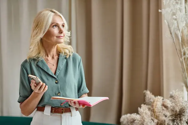 Bela mulher de meia idade com cabelo loiro segurando smartphone e segurando notebook na sala de estar — Fotografia de Stock