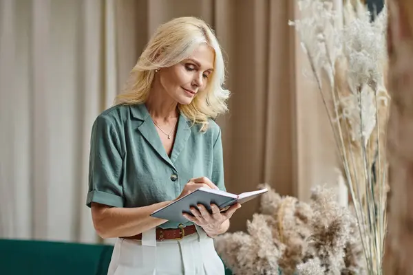 Beautiful middle aged businesswoman with blonde hair taking notes while holding notebook at home — Stock Photo
