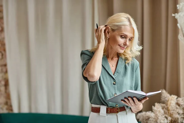 Séduisante femme d'affaires d'âge moyen avec des cheveux blonds tenant carnet tout en travaillant à la maison — Photo de stock