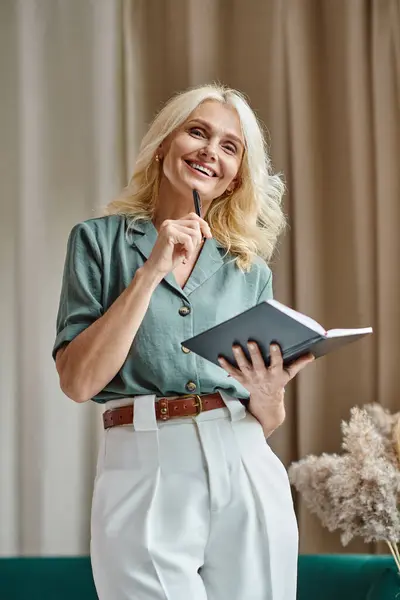 Joyeuse femme d'affaires d'âge moyen avec des cheveux blonds tenant un cahier et un stylo tout en travaillant à la maison — Photo de stock