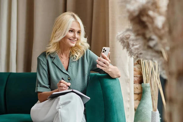 Fröhliche Geschäftsfrau mittleren Alters mit blonden Haaren, die in Notizbuch schreibt und zu Hause das Smartphone benutzt — Stockfoto