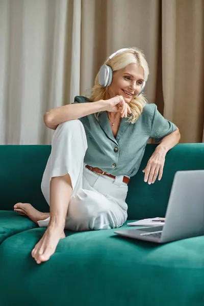 Heureuse femme d'âge moyen dans les écouteurs sans fil assis sur le canapé et en utilisant un ordinateur portable, travail de la maison — Photo de stock
