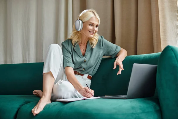 Joyeuse femme d'âge moyen dans les écouteurs sans fil assis sur le canapé et à l'aide d'un ordinateur portable, travail de la maison — Photo de stock