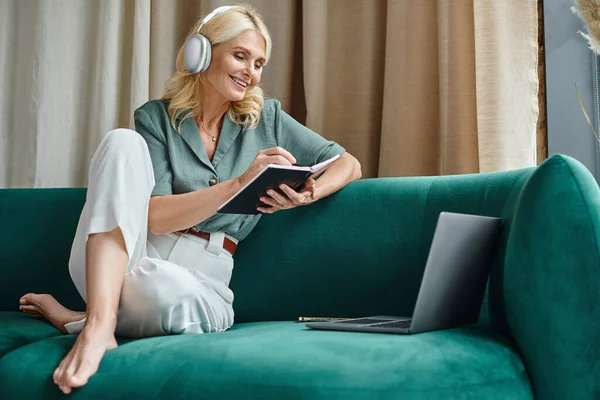Femme d'âge moyen positif dans les écouteurs sans fil assis sur le canapé près de l'ordinateur portable et portable — Photo de stock