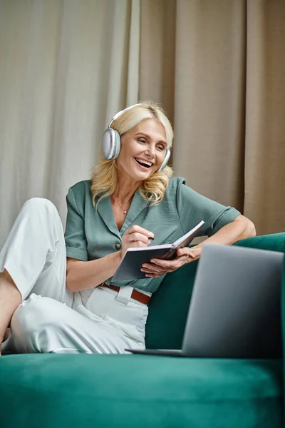 Mujer de mediana edad excitada en auriculares inalámbricos sentados en el sofá cerca de la computadora portátil y la escritura en portátil - foto de stock