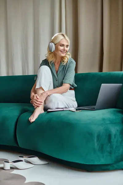 Mujer de mediana edad emocionada en auriculares inalámbricos sentados en el sofá cerca de la computadora portátil y webinar escuchar - foto de stock