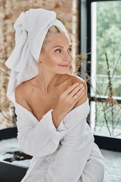 Heureux femme d'âge moyen avec serviette blanche sur la tête et peignoir appliquant gommage du corps sur l'épaule — Photo de stock