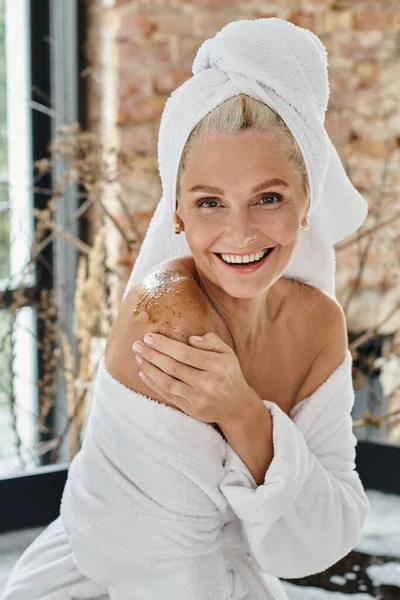 Mulher de meia idade alegre com toalha branca na cabeça e roupão aplicando esfoliação corporal, vertical — Fotografia de Stock