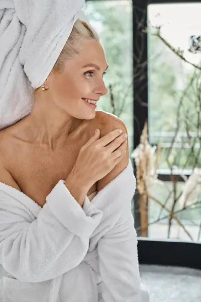 Smiling middle aged woman with white towel on head and bathrobe applying body scrub, vertical — Stock Photo