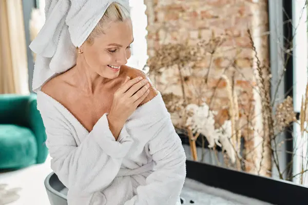 Joyeuse femme d'âge moyen avec serviette blanche sur la tête et peignoir appliquant gommage du corps, verticale — Photo de stock