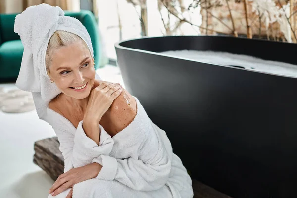 Feliz mulher de meia idade com toalha na cabeça e roupão de banho branco aplicando esfoliação corporal perto da banheira — Fotografia de Stock