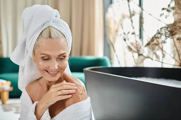 Happy middle aged woman in white bathrobe and with towel on head applying body scrub near bathtub — Stock Photo